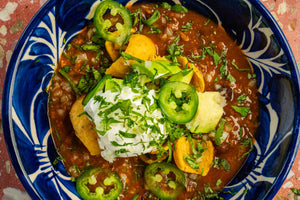 Bowl of chili topped with tortilla chips, sour cream, cilantro, jalapeños, and onion.